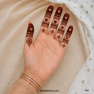 Front hand with dark fingertip mehndi, negative-space hearts, decorative finger bands, hanging dots and a small floral thumb motif.